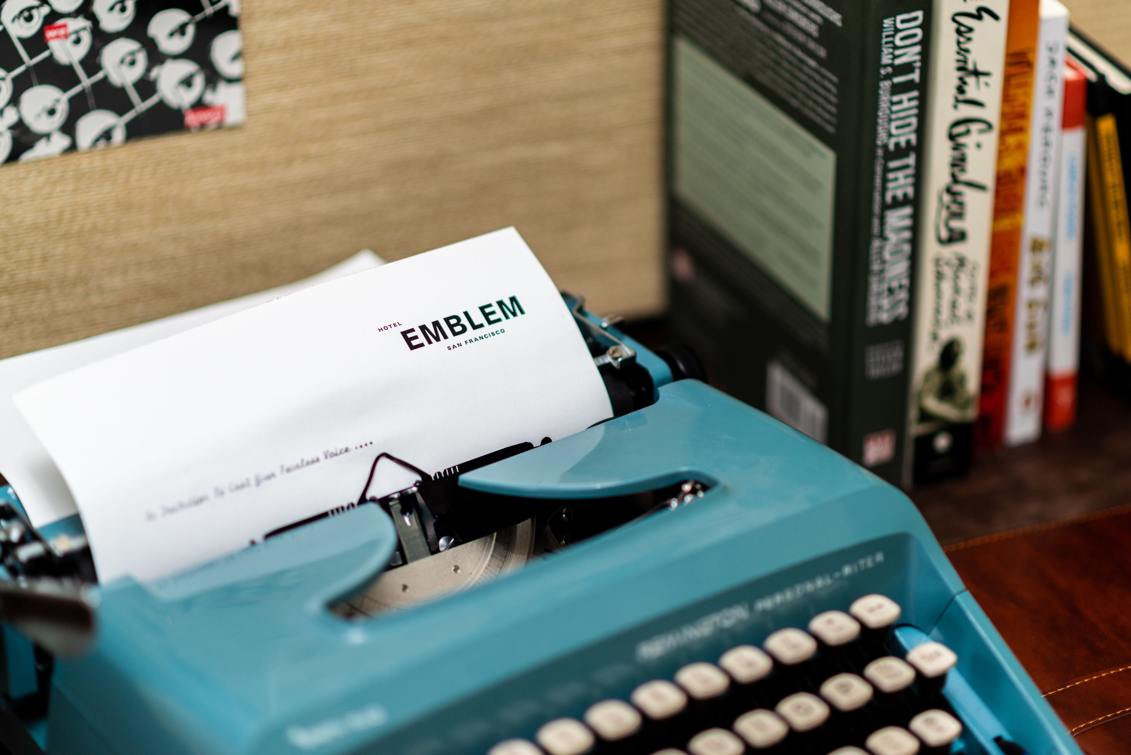 Une machine à écrire bleue, avec une feuille de papier sur laquelle est écrit le mot « EMBLEME », et un livre intitulé « NE CACHEZ PAS L’ARGENT » en arrière-plan.