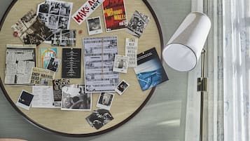 A room with a round bulletin board filled with photos and notes, a pink armchair with a cushion, a floor lamp, and a small side table with a wine glass.