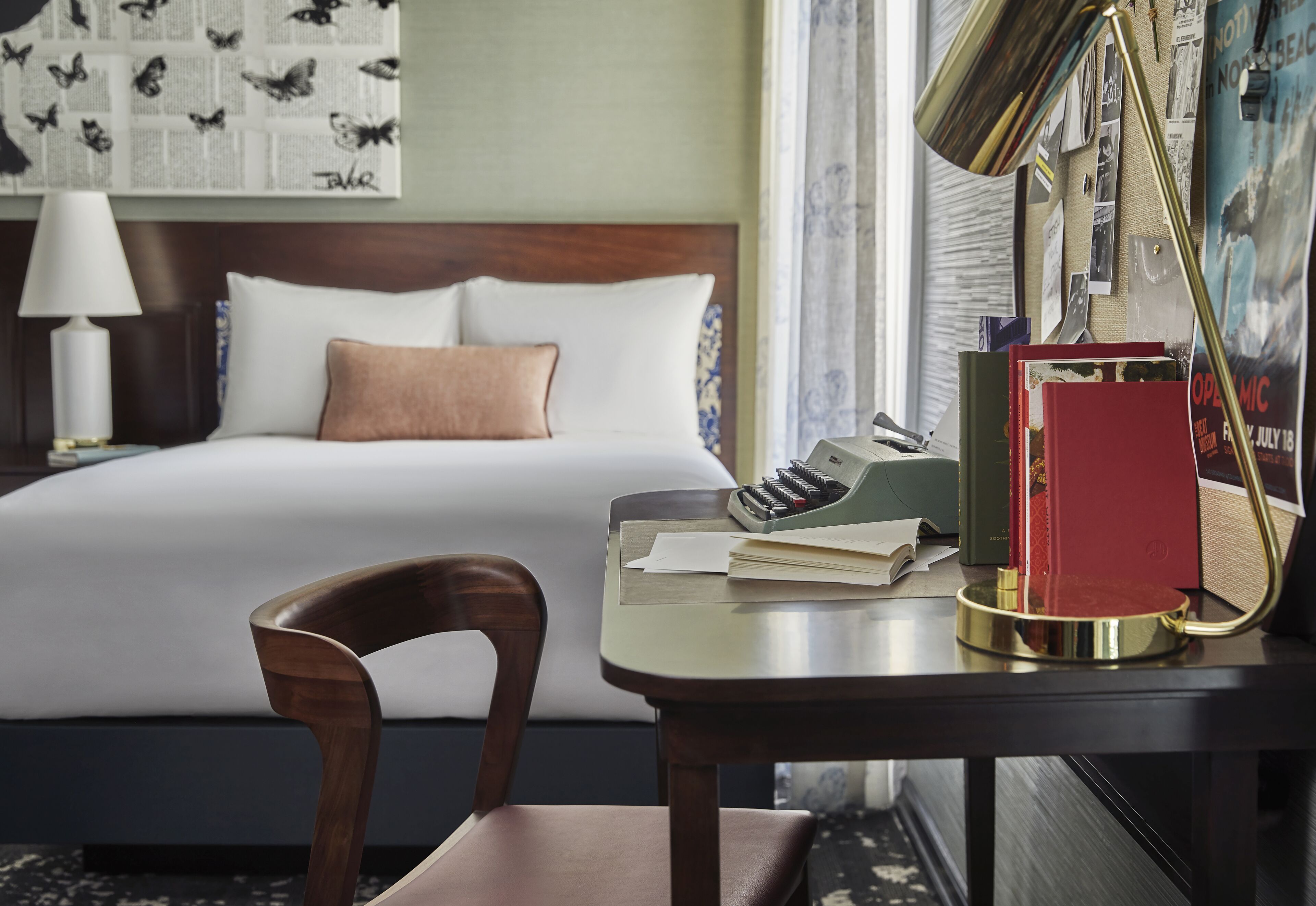 A hotel room with a neatly made bed, a desk with a lamp, a chair, and a bulletin board with papers and photos.