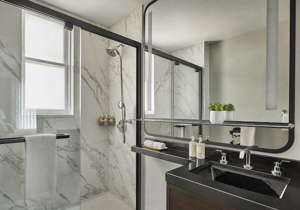 A modern bathroom with a glass shower enclosure, a marble-tiled wall, a black sink, and a mirror with a reflection of a plant.