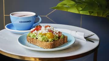 A breakfast setting with toast topped with avocado, tomato, and feta cheese, accompanied by a cup of coffee on a blue saucer.