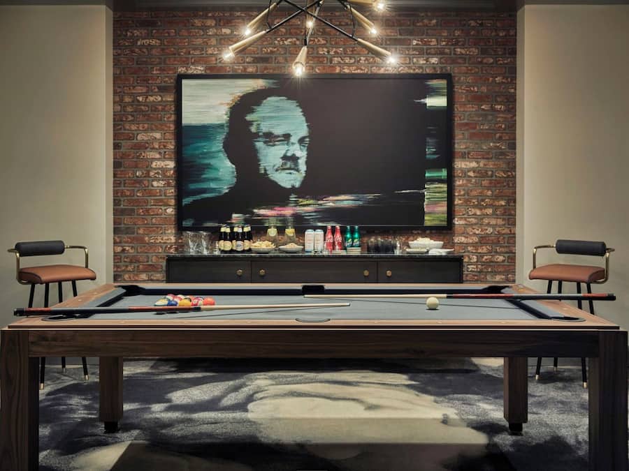 A pool table in a room with a brick wall and a large TV screen displaying a painting.