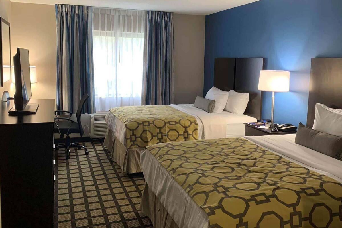 Ein Hotelzimmer mit zwei Betten, einem Schreibtisch mit Computer, einem Stuhl, einer Lampe und einem Fenster mit Vorhängen.