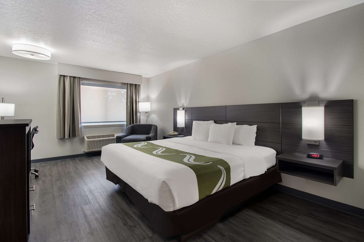 A hotel room with a large bed, a desk, a chair, a nightstand with a lamp, and a wall-mounted clock.