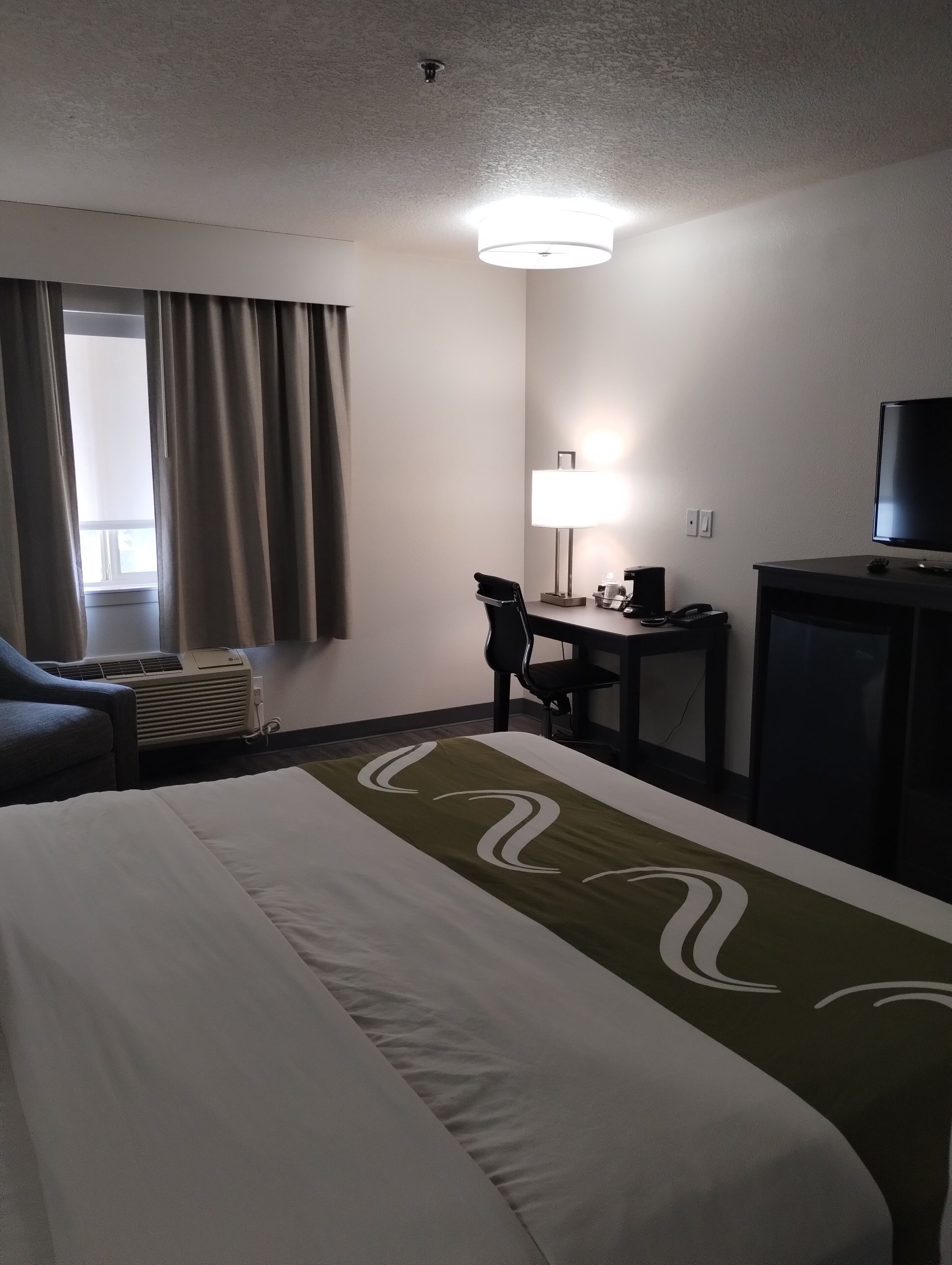 Habitación de hotel con cama, escritorio, silla, televisión y ventana con cortinas.