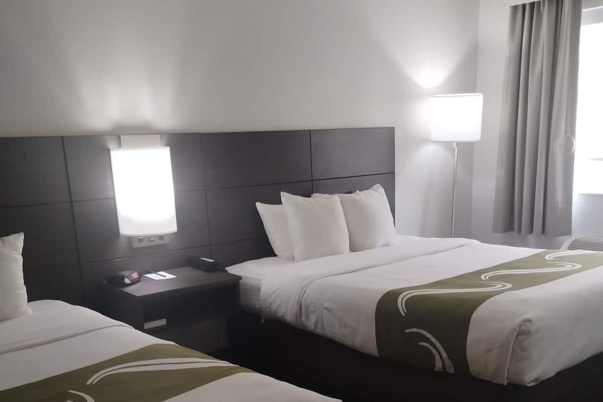 A hotel room with two beds, a bedside table with a lamp, and a window with curtains.