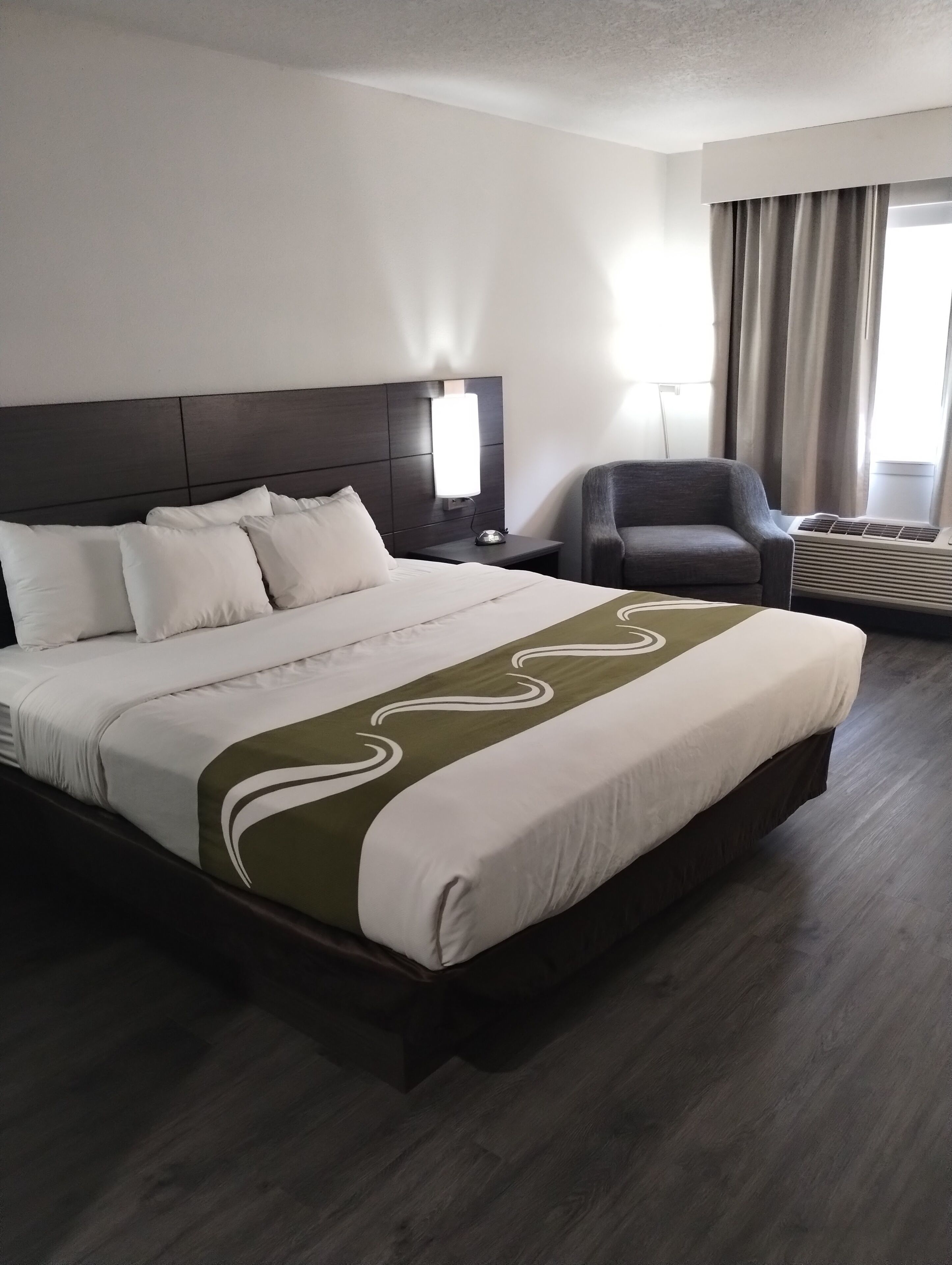 A hotel room with a large bed, a bedside table with a lamp, a chair, and an air conditioning unit.