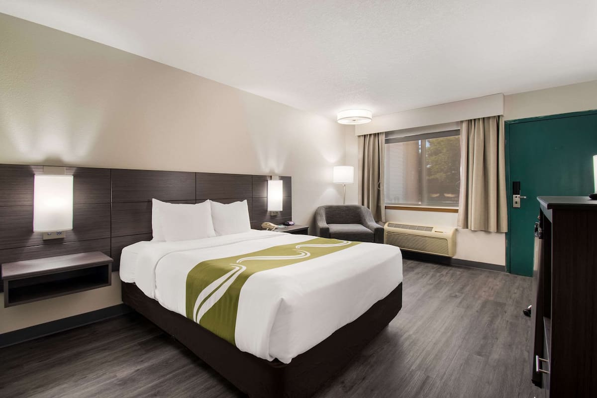 A hotel room with a large bed, bedside lamps, a chair, a desk, and a window with curtains.