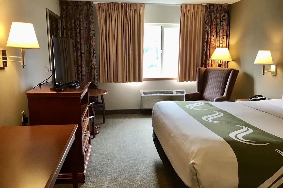 A hotel room with a bed, desk, chair, TV, air conditioning unit, and two lamps.
