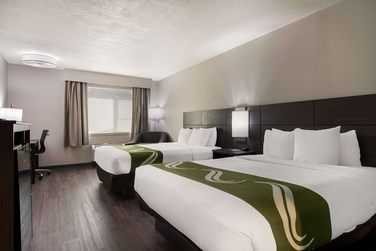 A hotel room with two beds, a desk, a chair, a lamp, and a window with curtains.