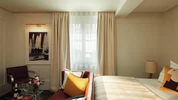 Een hotelkamer met een bed, een slaapbank, een glazen bijzettafeltje met een wijnfles en glazen, een vaas met bloemen en een schilderij aan de muur.