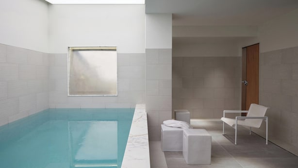 Una piscina interior moderna con vistas despejadas al cielo a través de un ventanal, rodeada de paredes de azulejos blancos y mobiliario minimalista.
