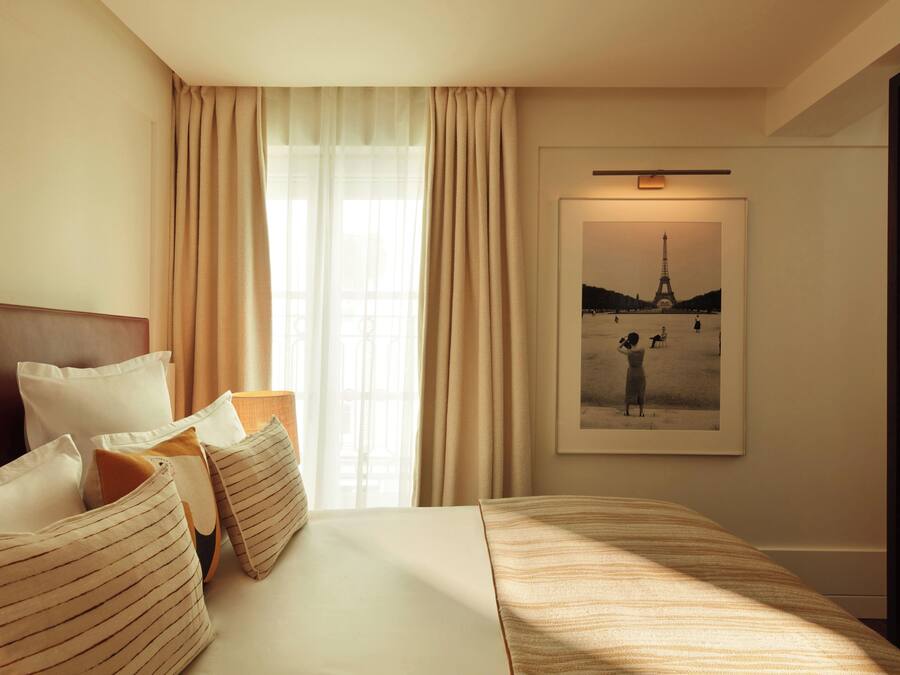 Une chambre d’hôtel avec un grand lit, des rideaux beiges, un tableau encadré représentant la Tour Eiffel et une tête de lit en bois.