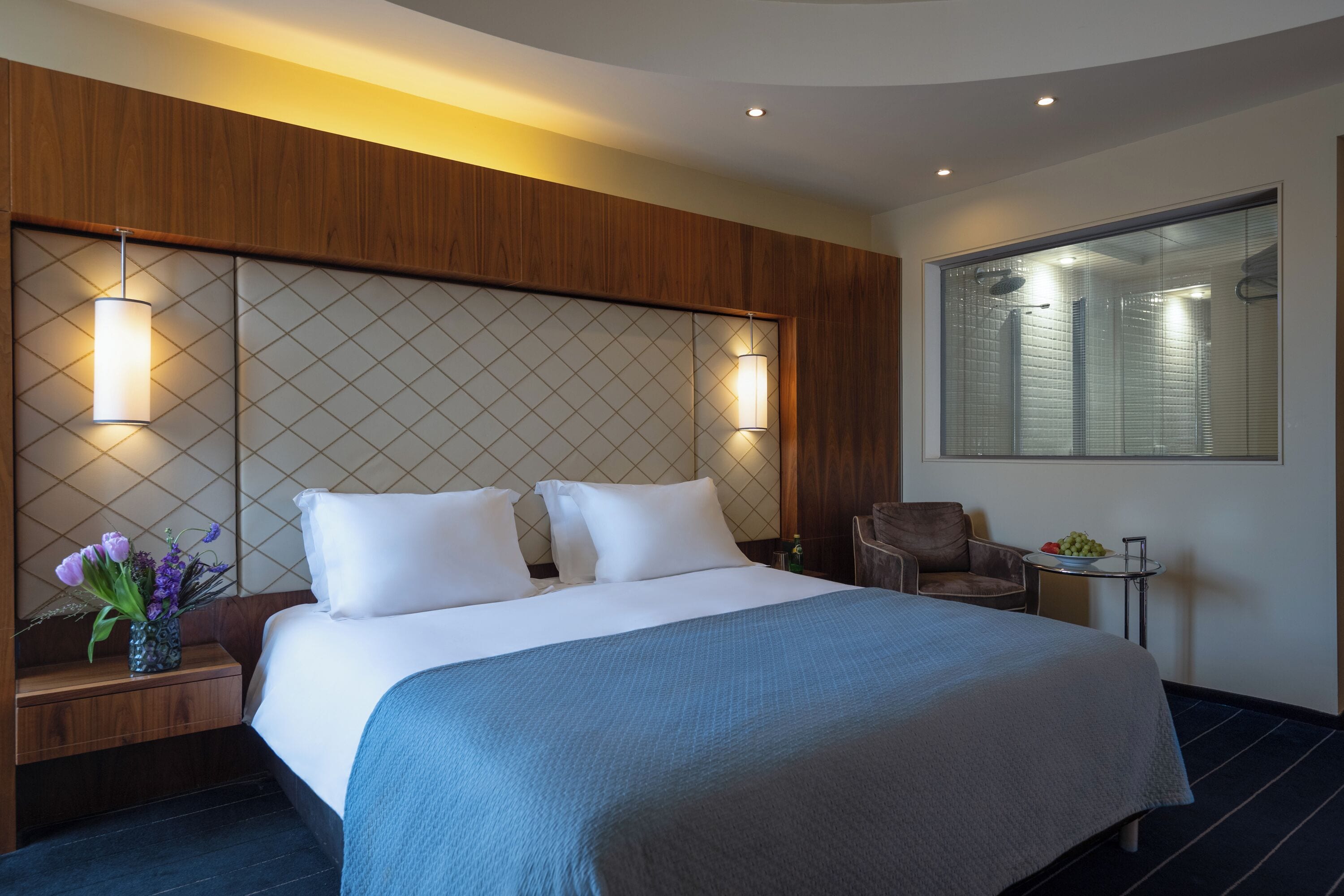 A hotel room with a large bed, a wooden headboard, two bedside lamps, a small table with a bowl of fruit, and a chair.