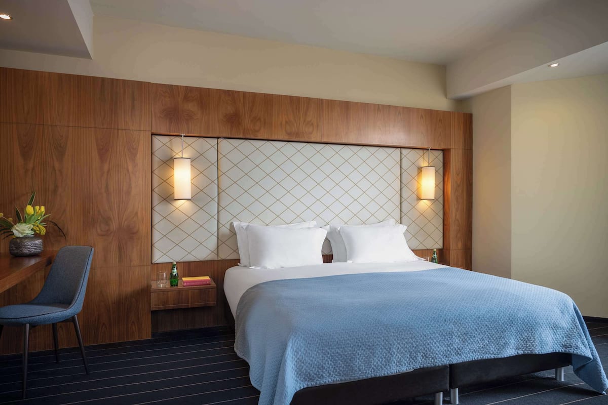 A hotel room with a large bed, a wooden headboard, two wall-mounted lamps, a small table with a chair, and a potted plant.