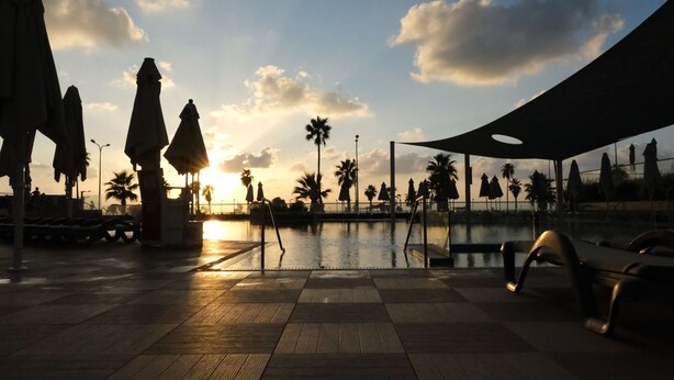 A poolside area with umbrellas, palm trees, and a sunset in the background. - Dan Panorama Tel Aviv (Tel Aviv)