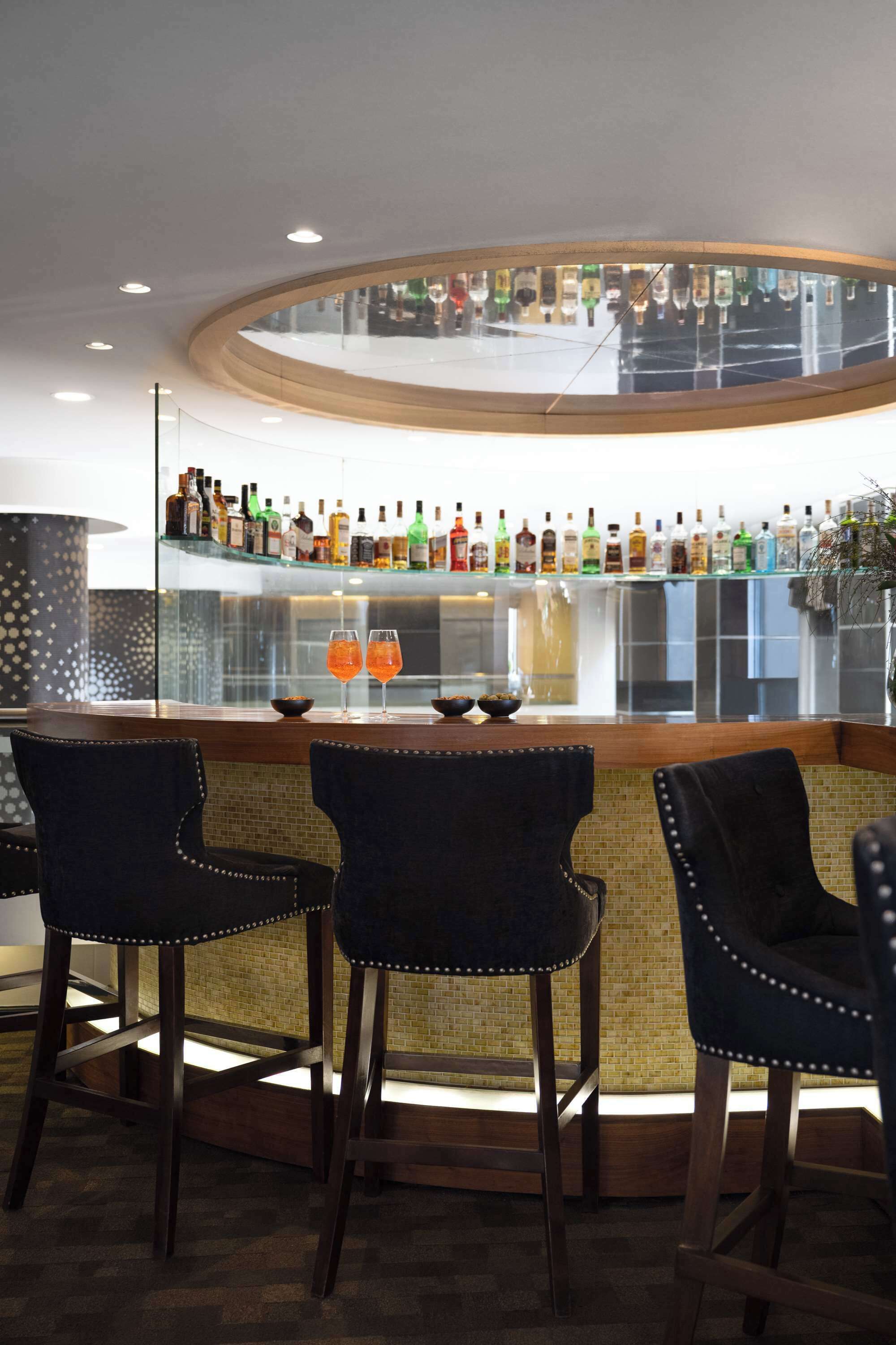 A bar area with a curved counter, three upholstered bar stools, and a variety of bottles on shelves behind the counter.