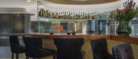 Zona de bar con barra circular, varias botellas de licor y tres taburetes.