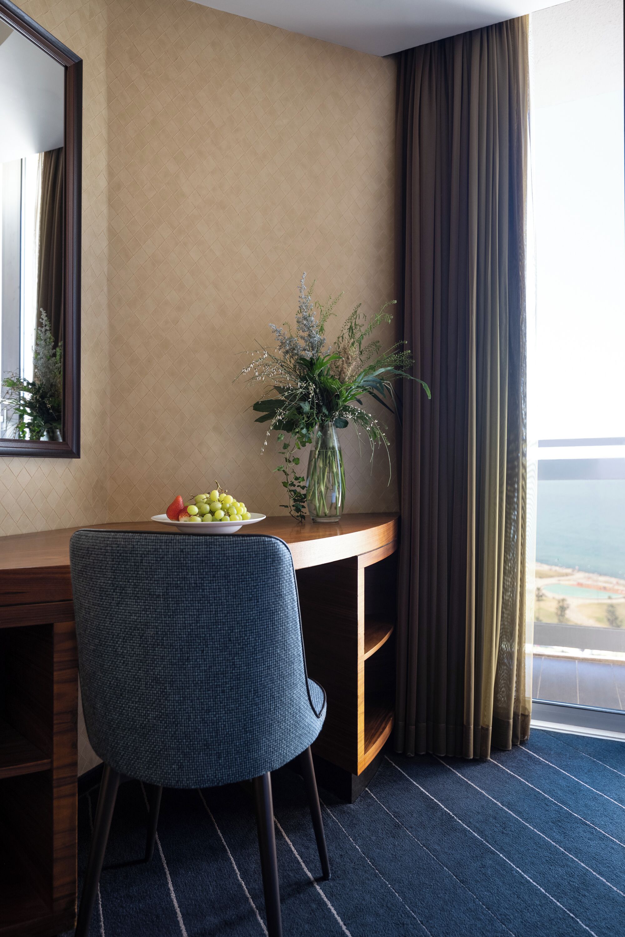 A hotel room with a desk, a chair, a vase with flowers, and a view of the sea.