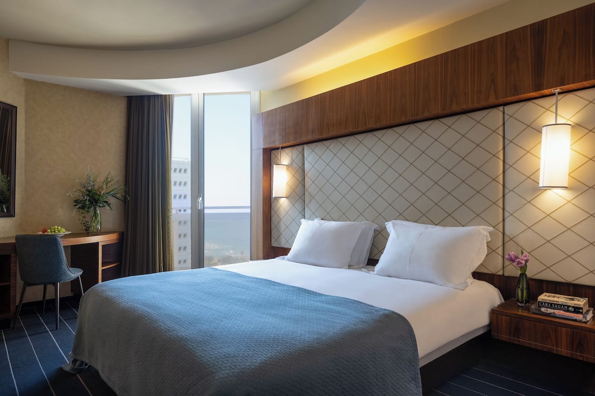 A hotel room with a large bed, a desk with a chair, a nightstand with books and a vase, and a view of the city through a large window.