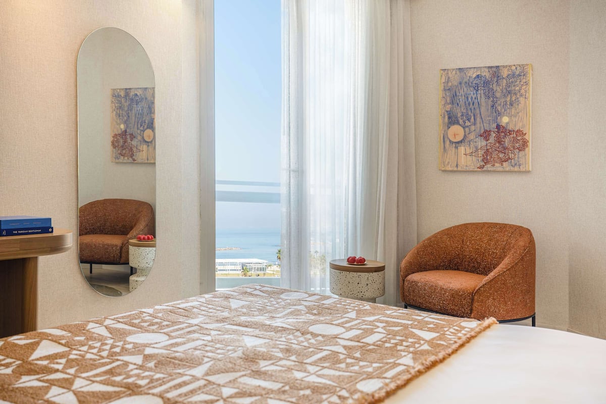 A hotel room with a bed, two armchairs, a mirror, a small table with a book and fruit, and artwork on the walls.