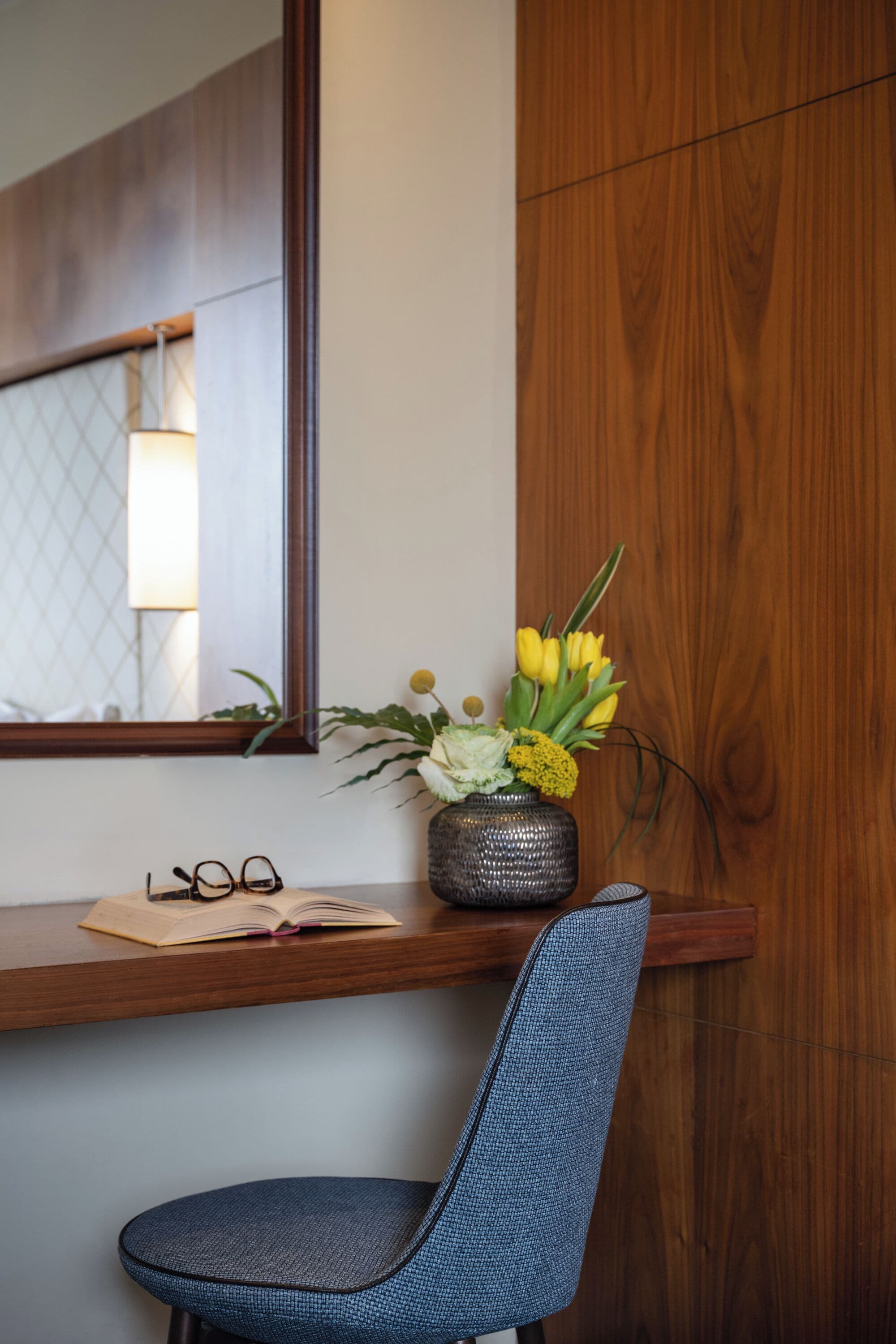 A wooden desk with a vase of flowers, a pair of glasses, and an open book.