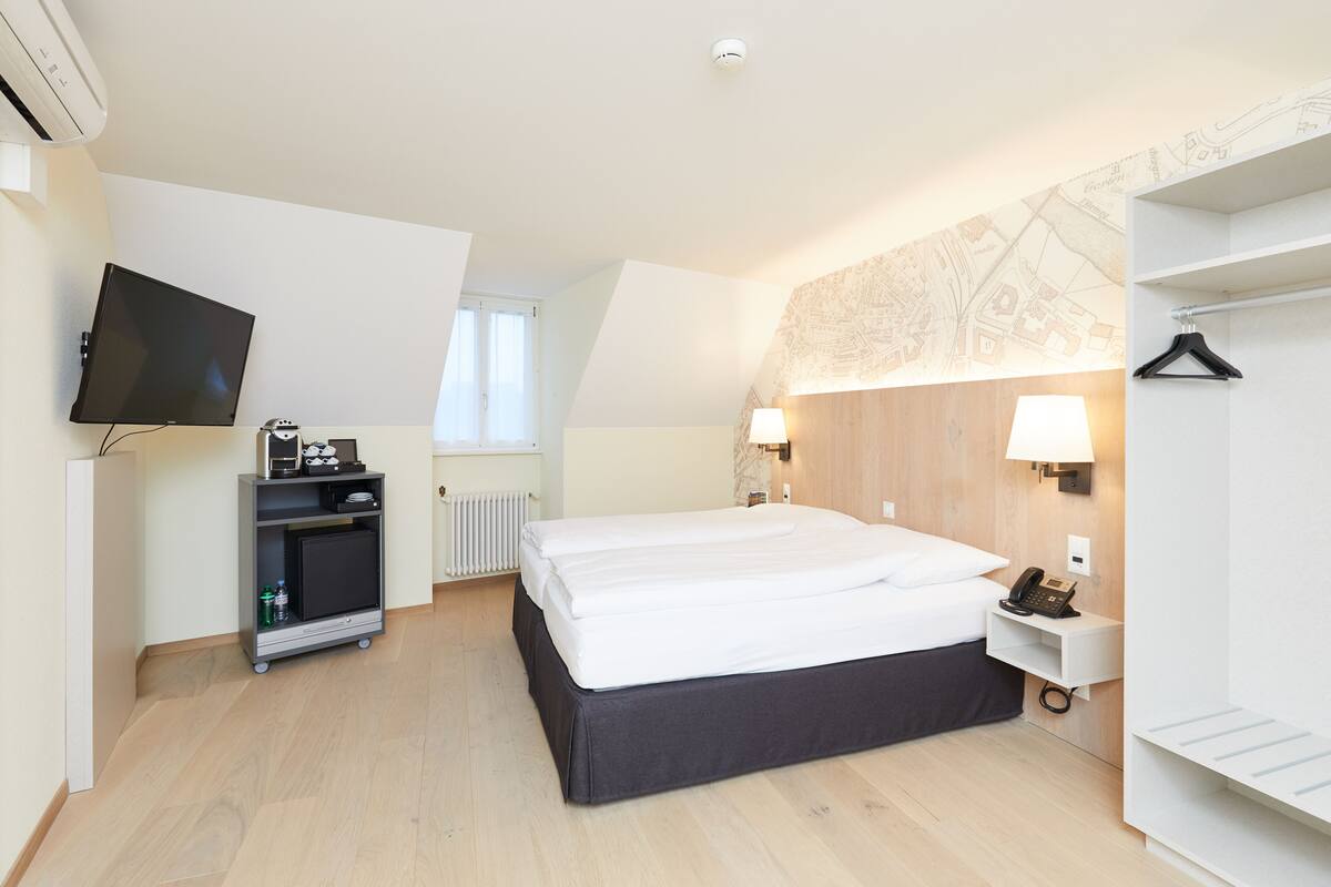 A hotel room with a bed, bedside table, lamp, TV, and a cart with items.