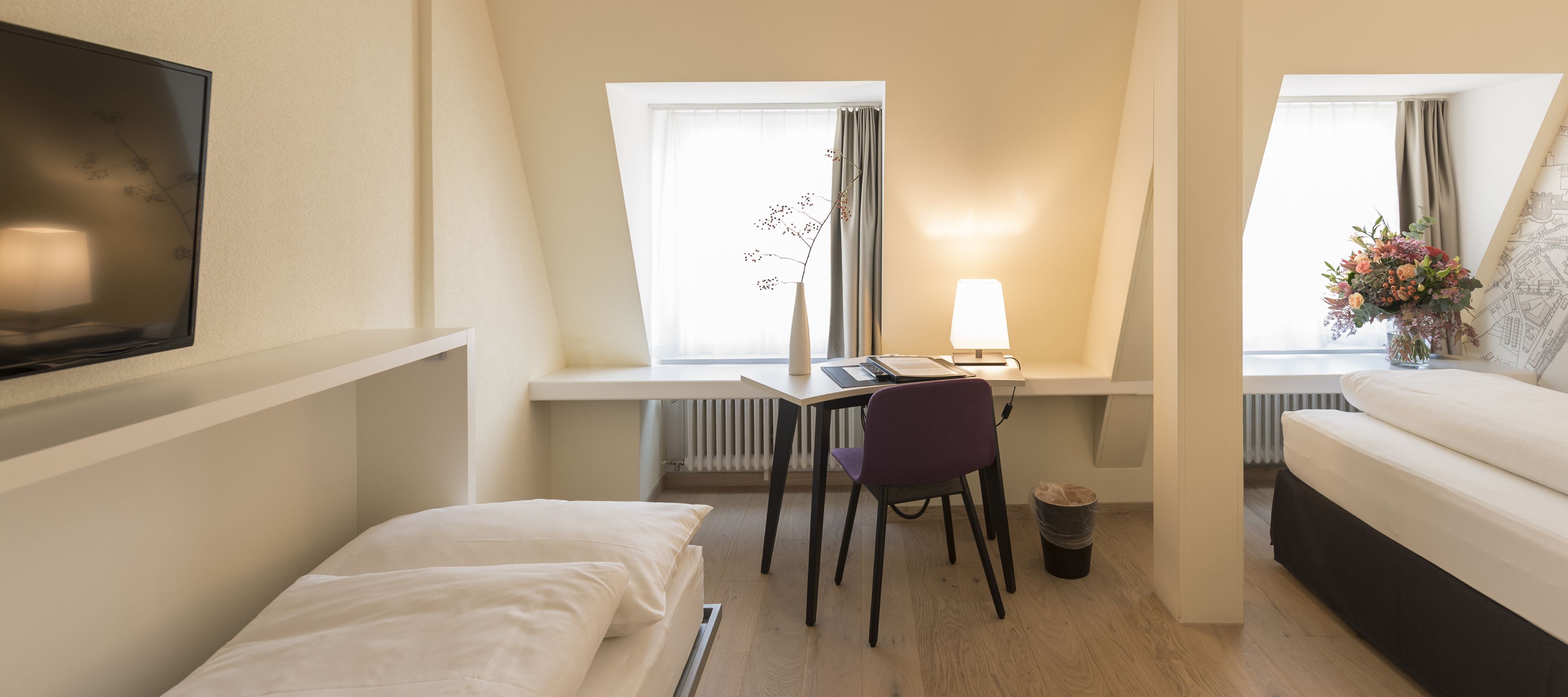 A compact hotel room with a bed, a desk with a chair, a TV mounted on the wall, and a window with curtains.
