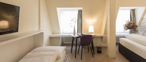 A compact hotel room with a bed, a desk with a chair, a TV mounted on the wall, and a window with curtains.