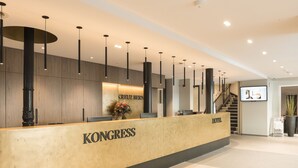 Un comptoir d’accueil d’hôtel avec un comptoir en bois, l’inscription « KONGRESS » bien visible, et un escalier menant aux étages supérieurs.