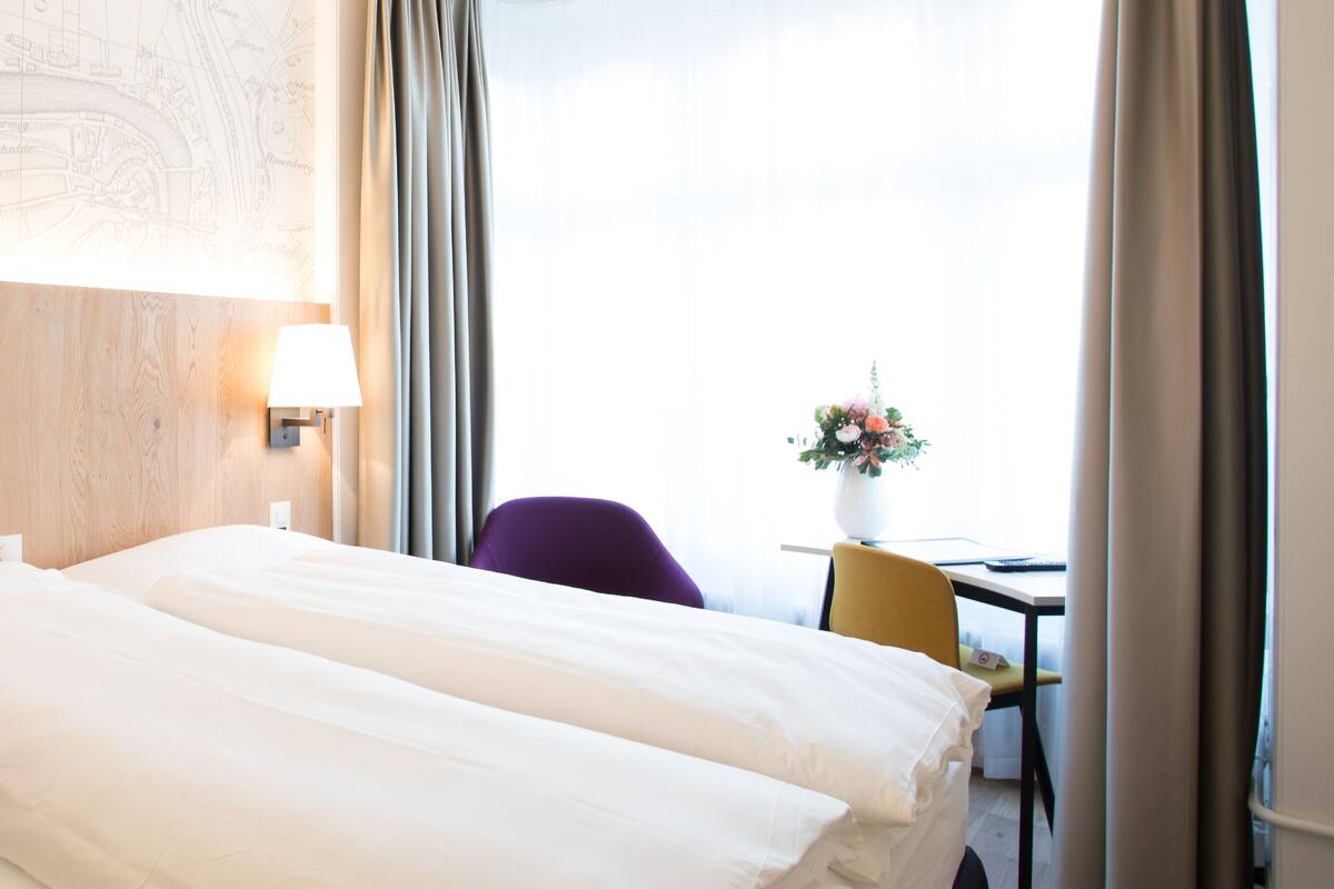 A hotel room with a bed, a desk, a chair, a lamp, and a map on the wall.