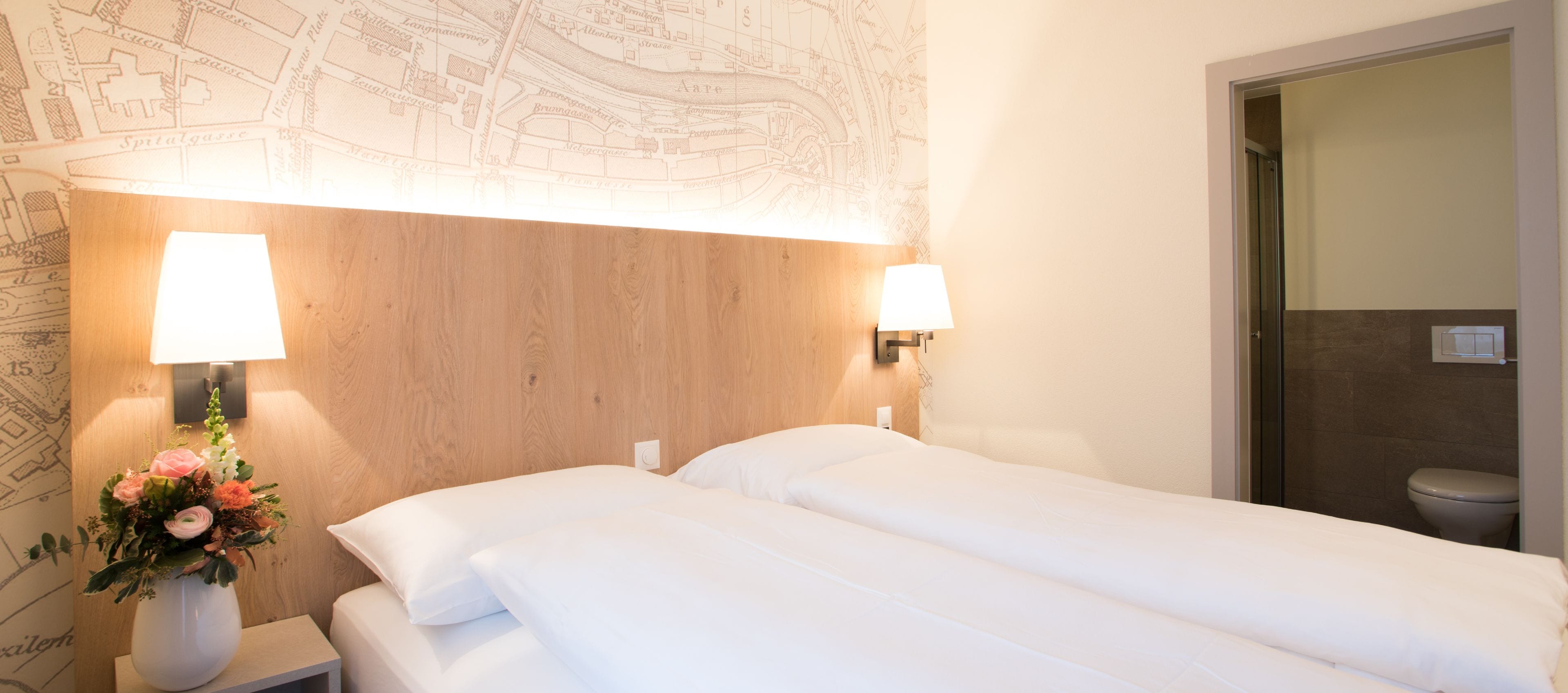 Une chambre d’hôtel avec deux lits, une tête de lit en bois, une carte accrochée au mur et un vase rempli de fleurs.