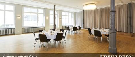 Un salón amplio con suelos de madera y mesas blancas dispuestas para comer, columnas y un gran ventanal con vistas.