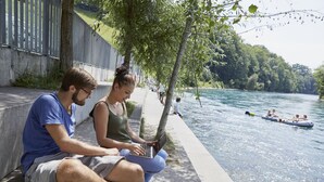 Deux personnes sont assises sur une promenade au bord de la rivière, et on aperçoit un bateau sur l’eau au loin.
