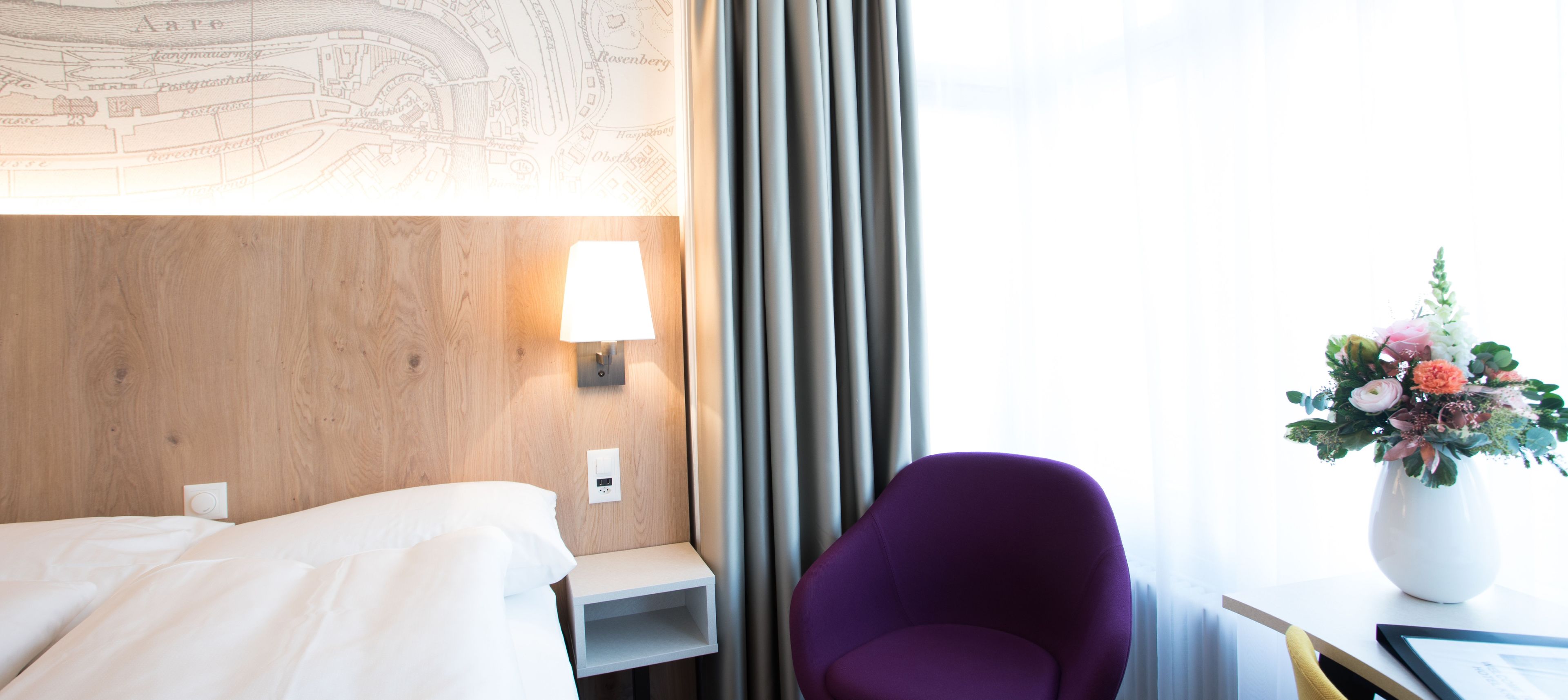 A hotel room with a bed, a nightstand with a lamp, a purple chair, and a window with curtains.