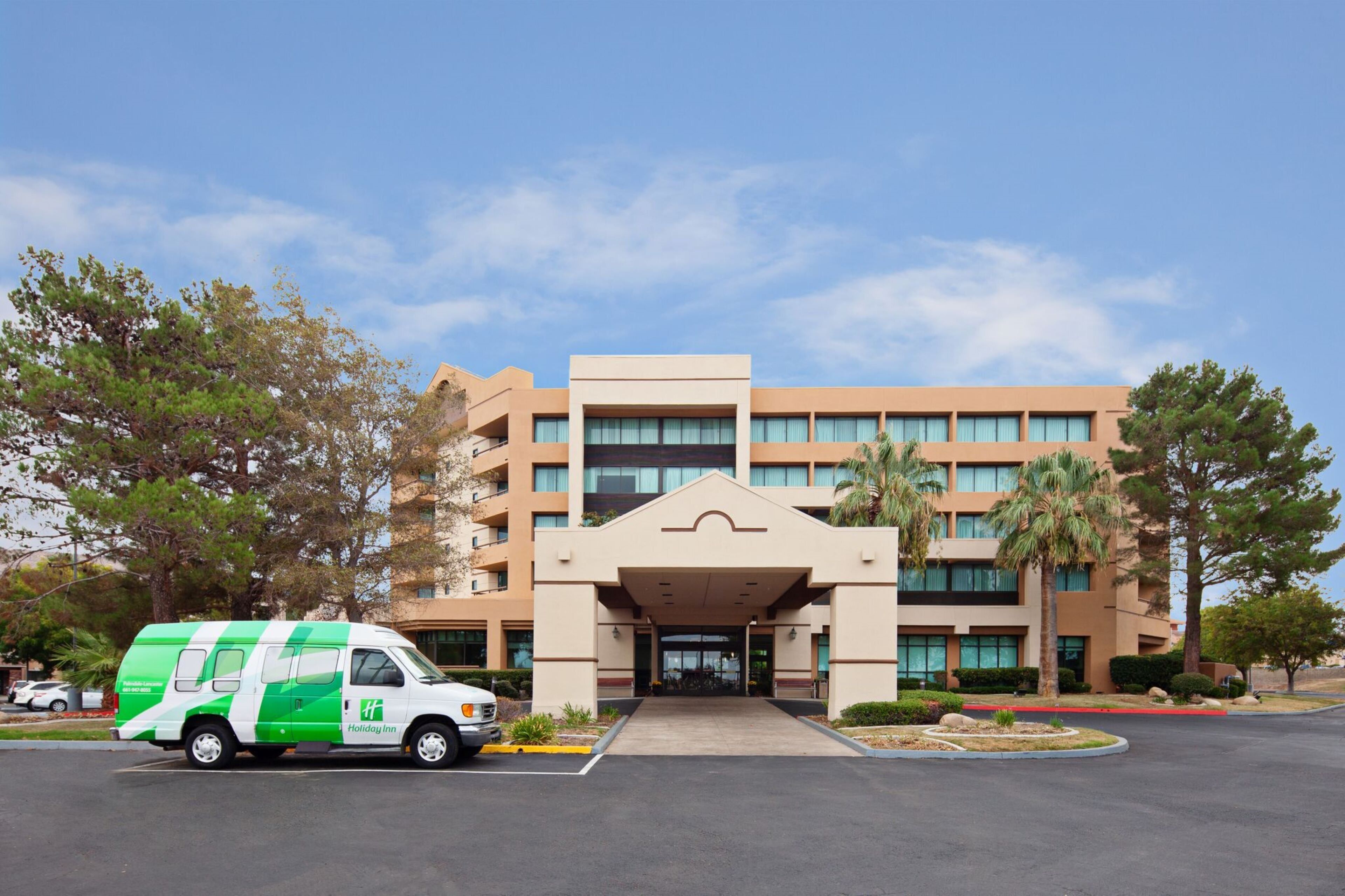 A multi-story hotel with a central entrance, a parking lot, and a green and white shuttle service.