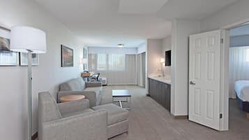 A hotel room with a sofa, a small table, a desk with a chair, a TV, and a door leading to a bathroom.