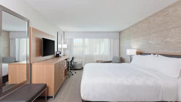 A hotel room with a large bed, a desk with a flat-screen TV, a chair, a desk lamp, and a large mirror.