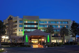 Un hotel de varios pisos con una entrada central, rodeado de áreas ajardinadas y palmeras.
