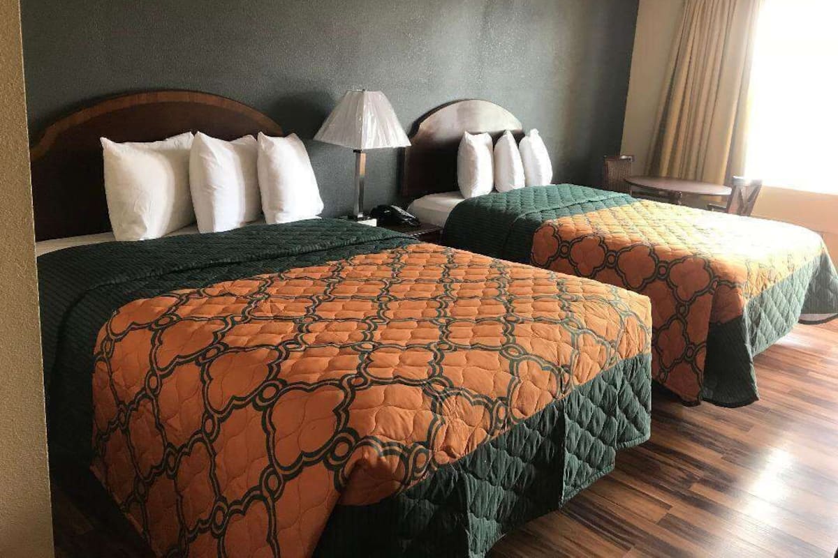 A hotel room with two beds, a nightstand, a lamp, and a window with curtains.