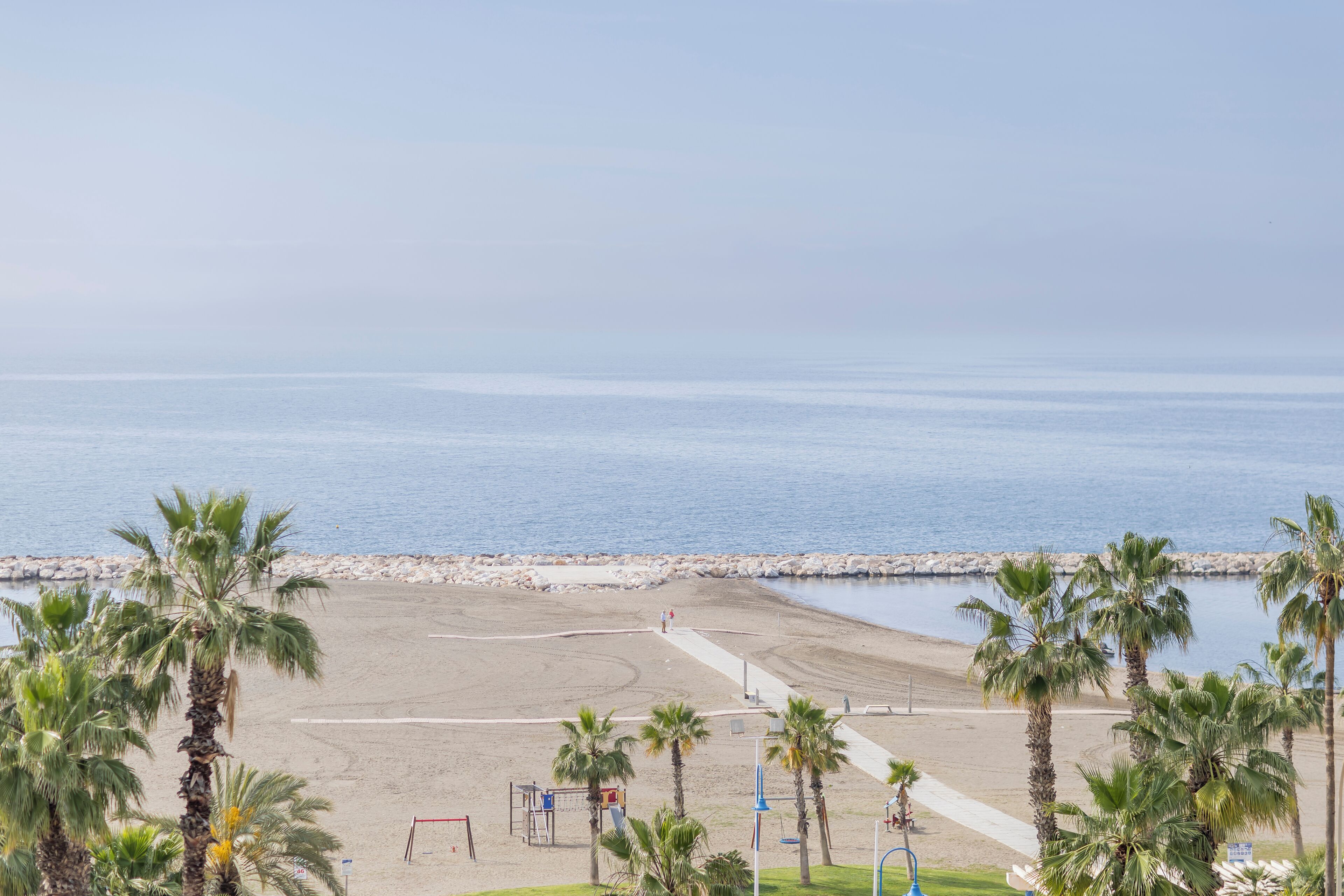 A beach with a volleyball court, palm trees, and a rocky breakwater.