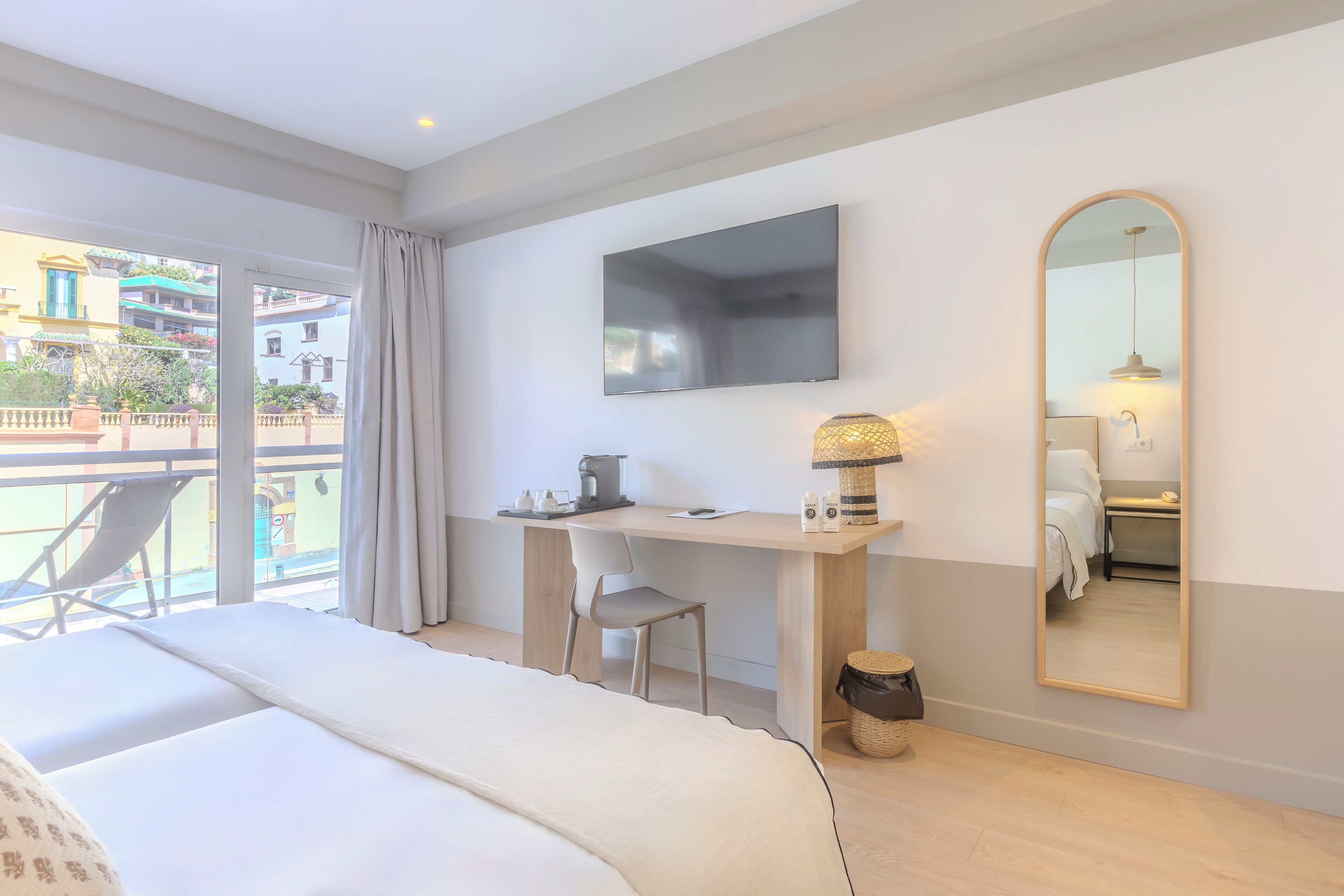A hotel room with a bed, a desk, a chair, a TV, a mirror, and a balcony with a view of buildings.