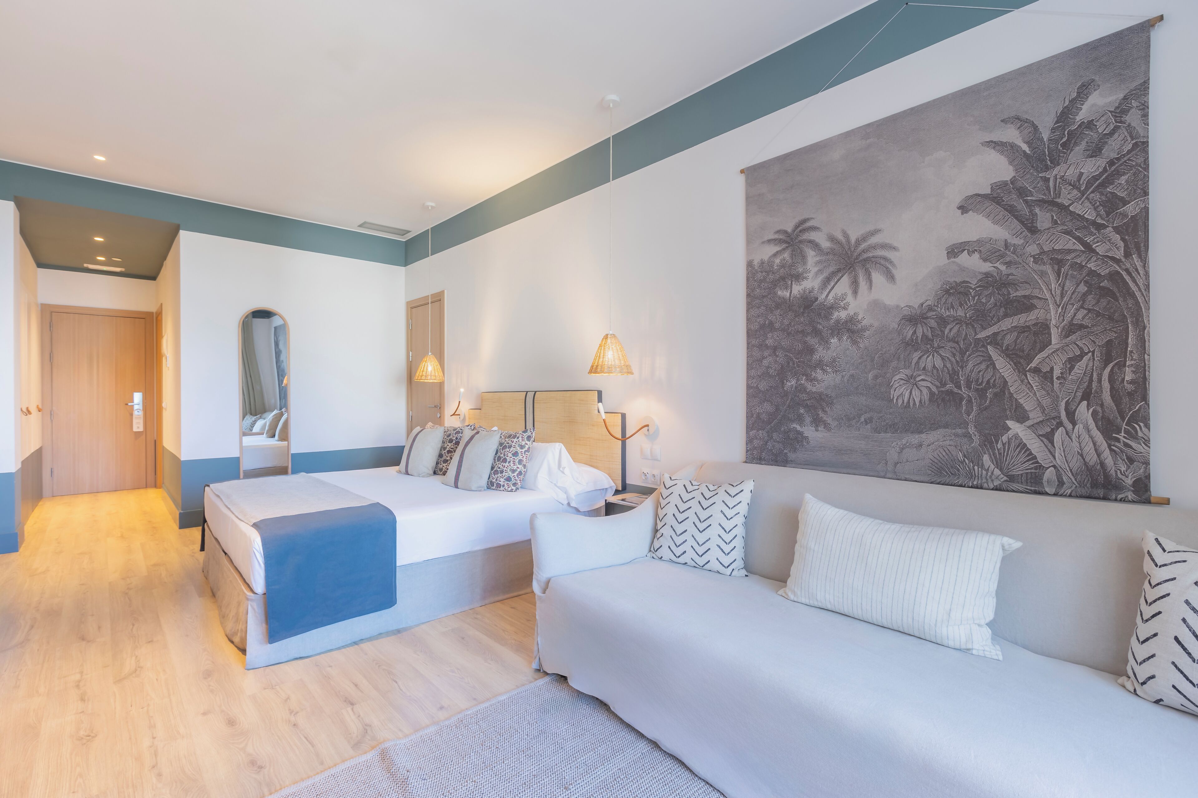 A modern hotel room with a large bed, a sofa, a desk, and a large tropical mural on the wall.
