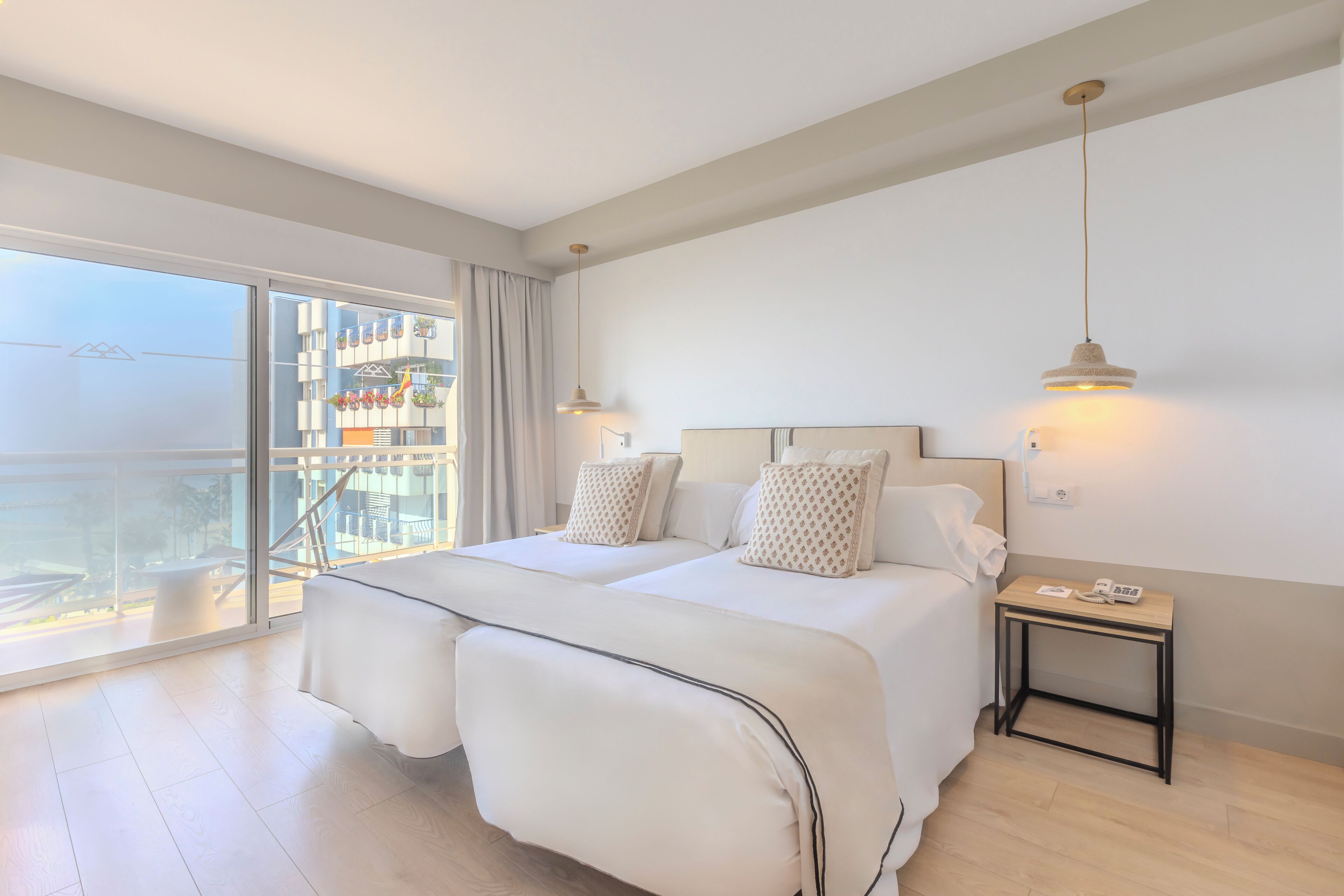 A hotel room with a large balcony, two beds, a bedside table with a phone, and a hanging light fixture.