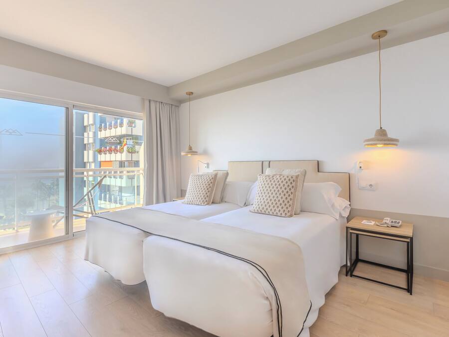 A hotel room with a large balcony, two beds, a bedside table with a phone, and a hanging light fixture.