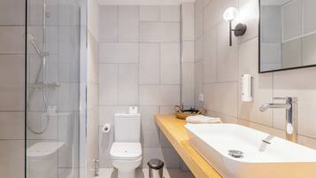Un baño moderno con ducha envuelta en vidrio, inodoro blanco, tocador de madera con un lavamanos grande y un espejo.