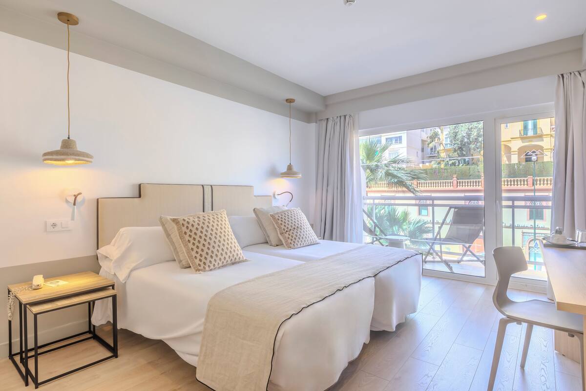 A hotel room with two beds, a small table, a chair, and a balcony with a view of the city.