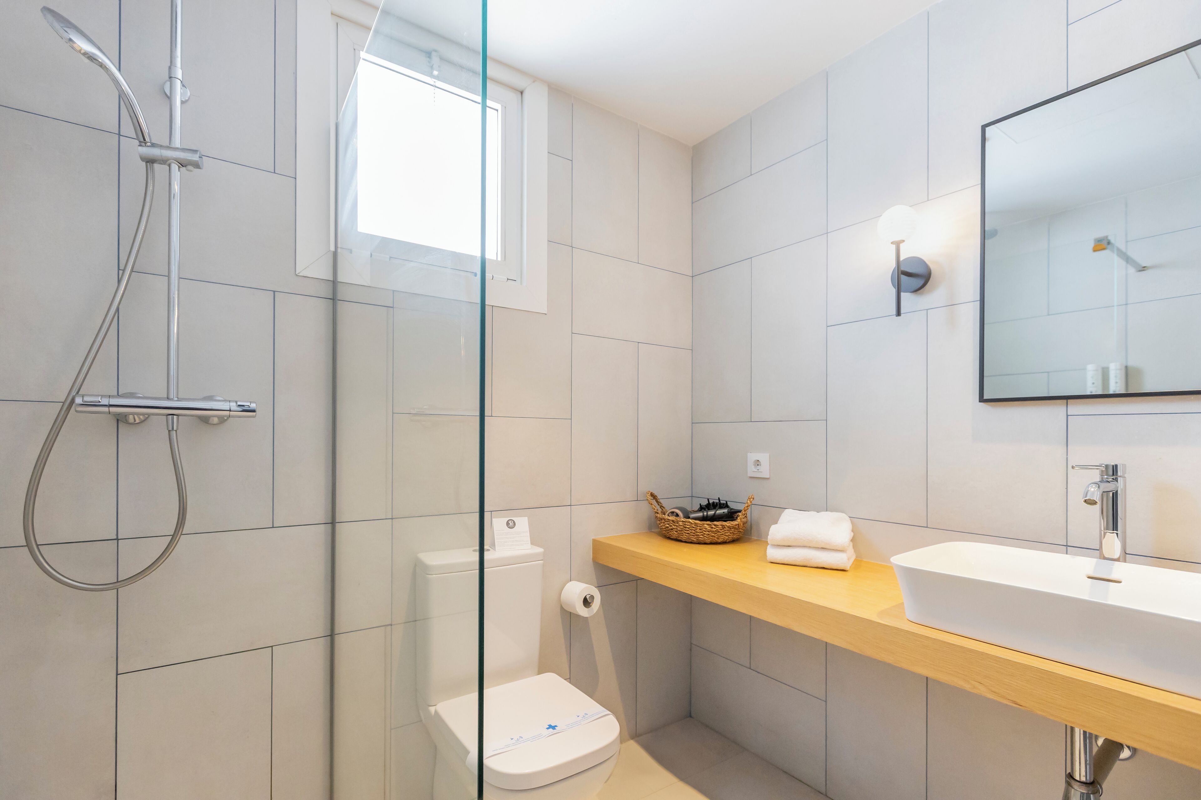 Une salle de bain moderne dotée d’une douche à parois de verre, de toilettes, d’un lavabo avec un miroir et d’une fenêtre qui laisse entrer la lumière naturelle.