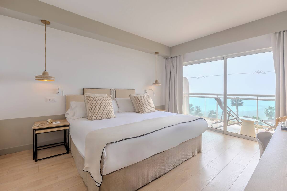 A modern hotel room with a large bed, bedside tables, a desk, and a balcony with a view of palm trees.