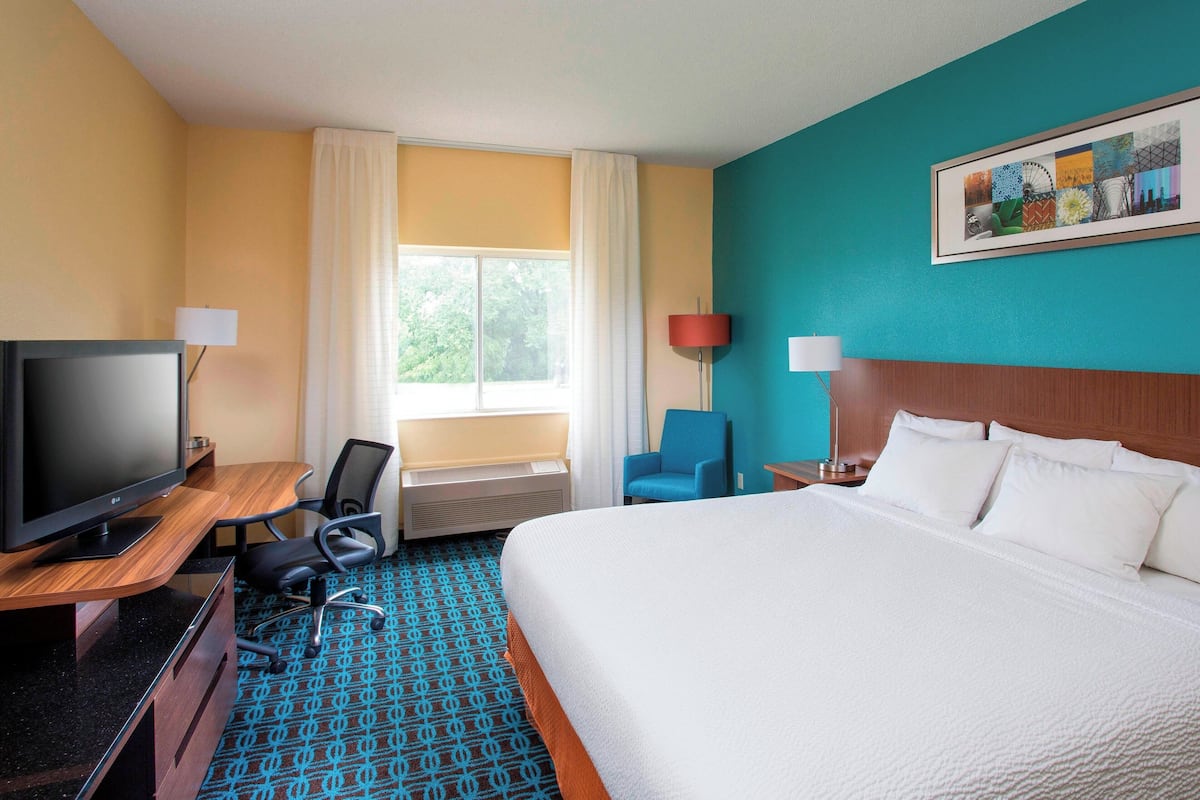 A hotel room with a large bed, a desk with a TV, a chair, a window with curtains, and a painting on the wall.